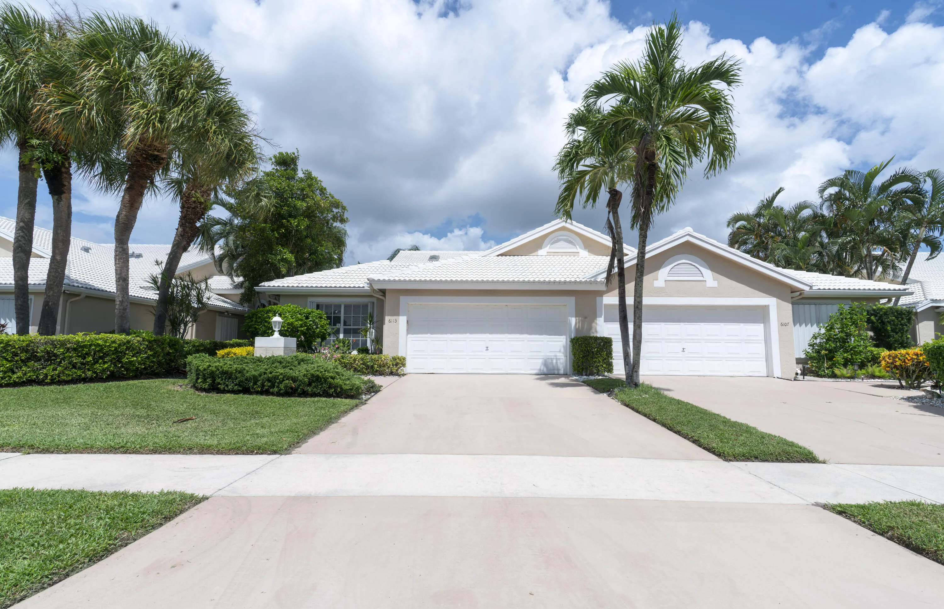 Exterior featuring a white tile roof, dual garage doors, a concrete driveway, established palm trees, and manicured landscaping