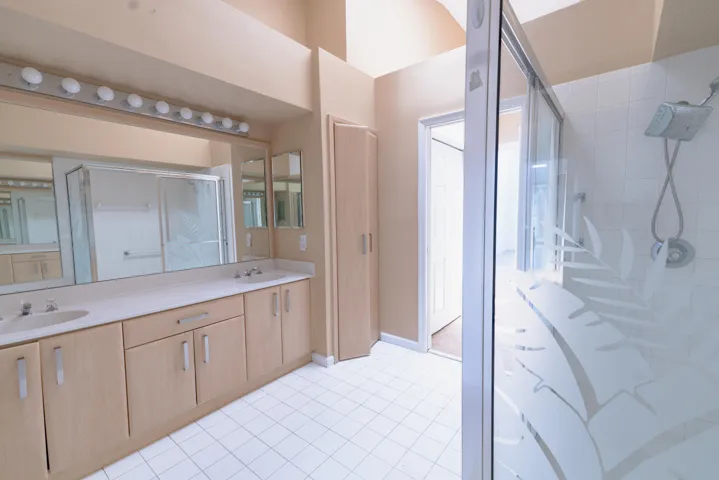 Dual vanity with integrated sinks and ample countertop space, extensive wood-finish cabinetry, vaulted ceiling, tile flooring, and a glass-enclosed shower with a handheld showerhead