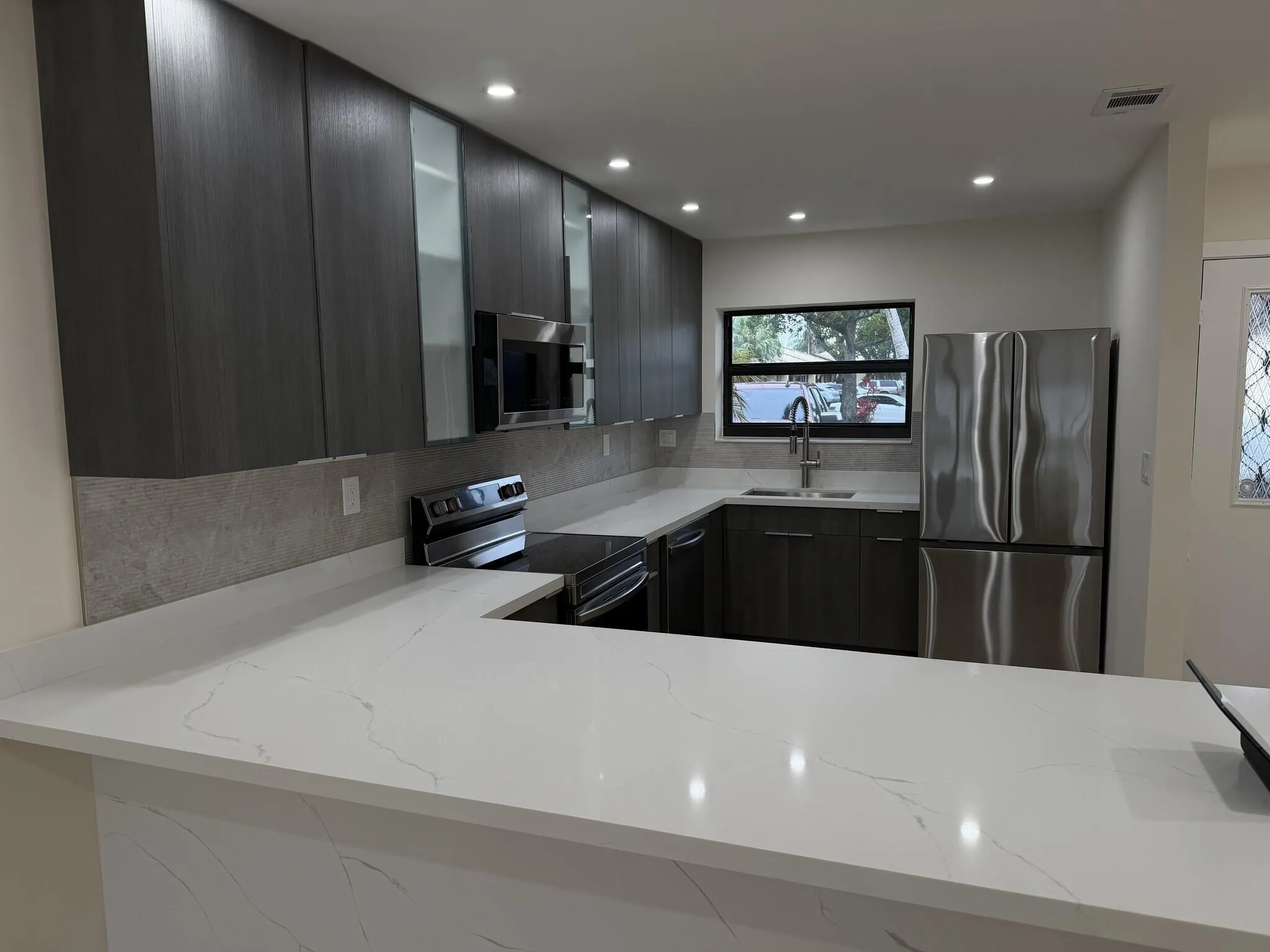 Contemporary kitchen featuring dark wood-finish cabinetry, light-toned countertops, a light-toned backsplash, stainless steel appliances, and recessed lighting