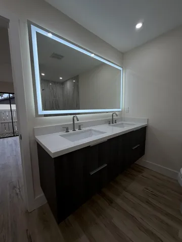 Floating double vanity with integrated sinks, white countertop, illuminated mirror, recessed lighting, and wood-finish flooring