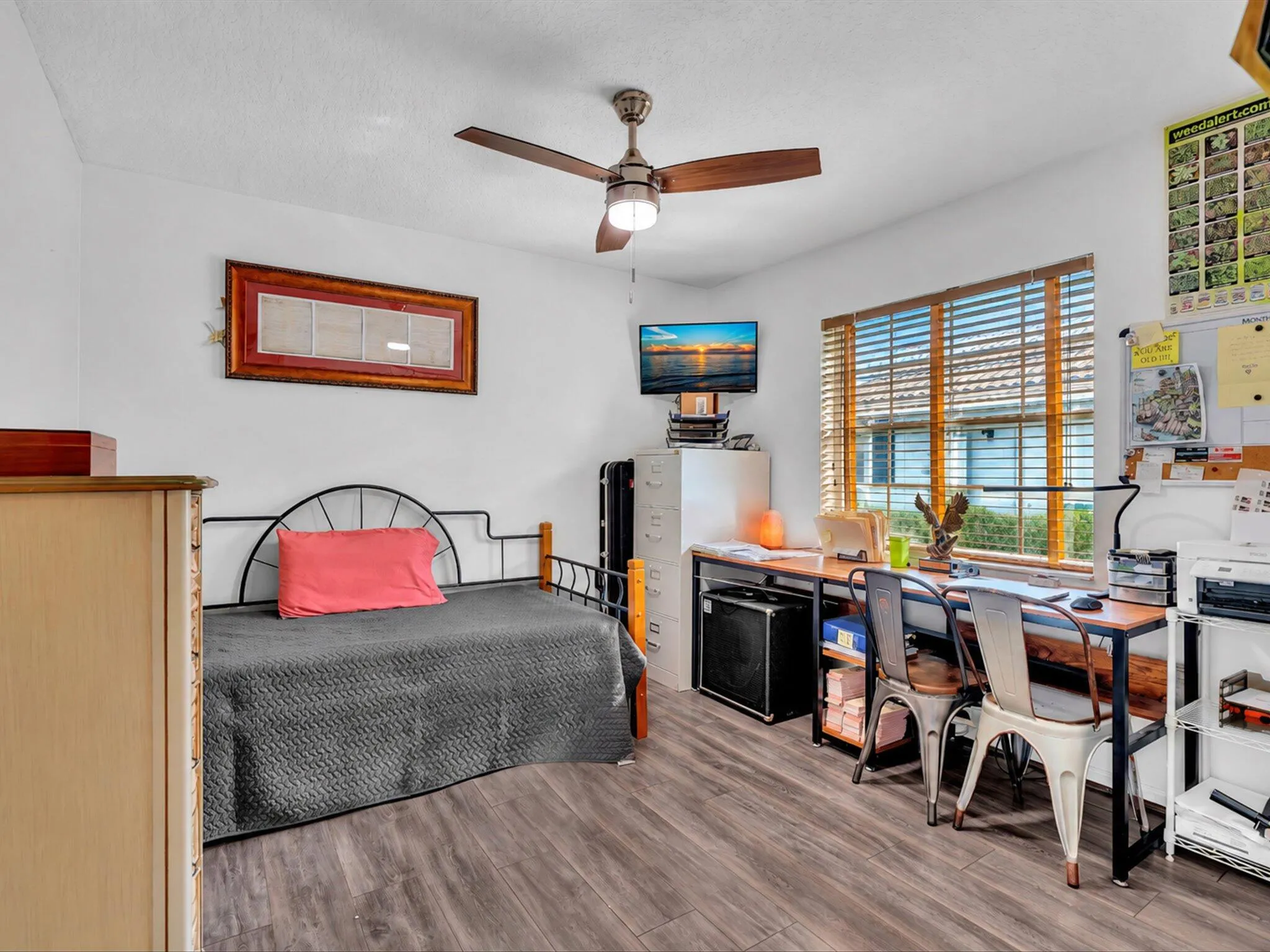 Wood-finish flooring, ceiling fan with integrated lighting, window with horizontal blinds, built-in desk with metal frame, and white walls