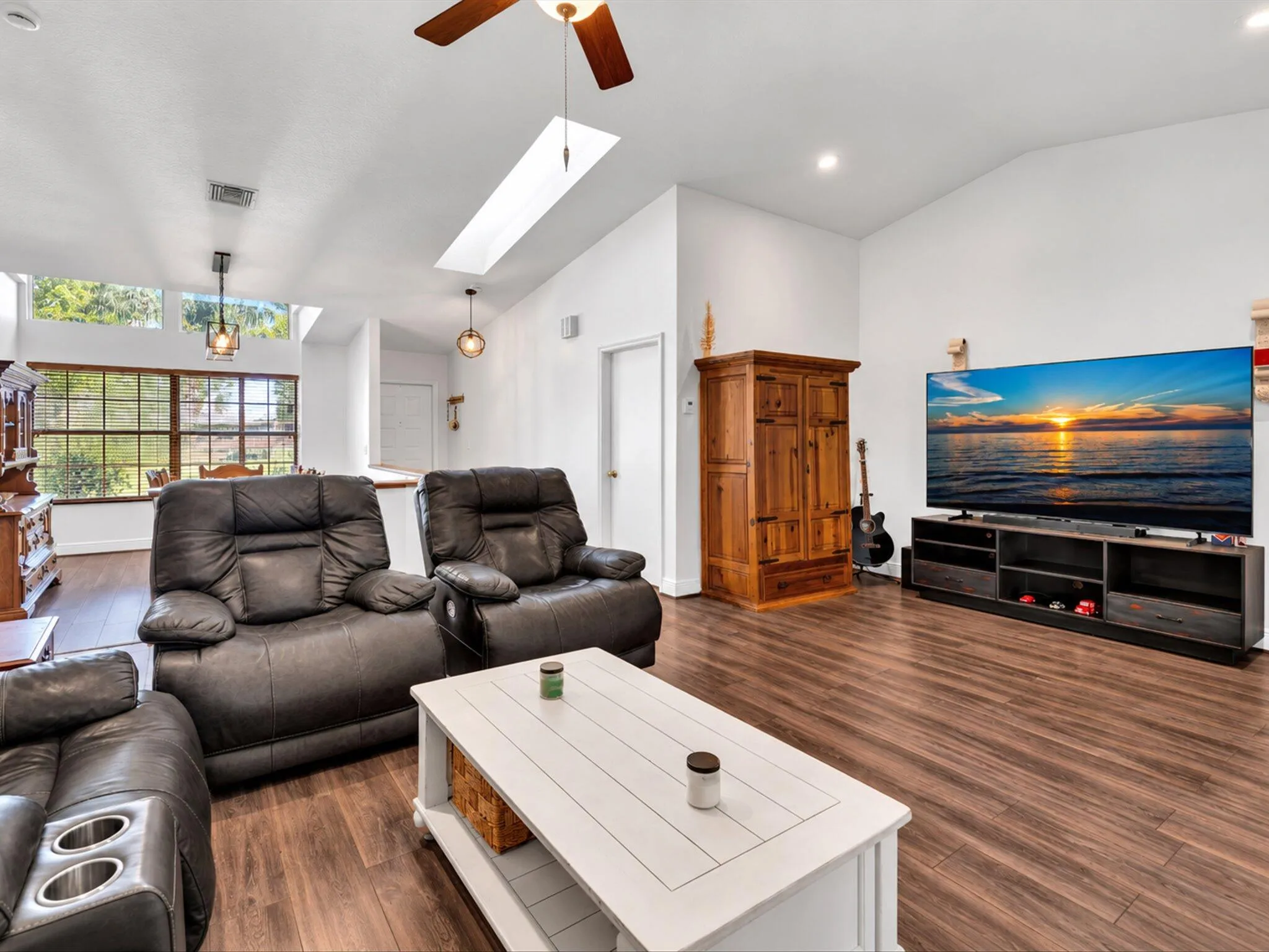 Vaulted ceiling with skylight, wood-finish flooring, ceiling fan, recessed lighting, and large windows with exterior foliage