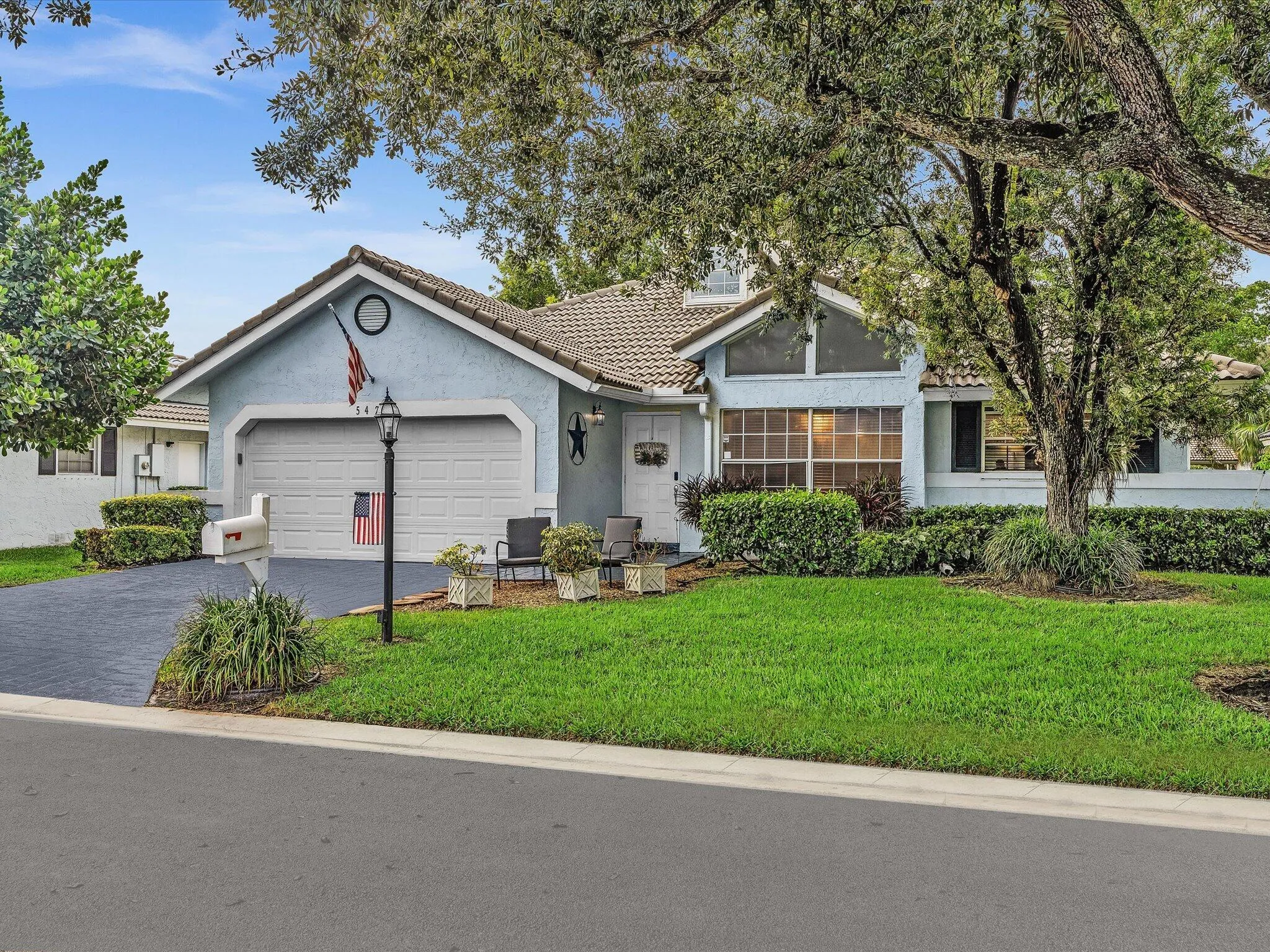 Residential property featuring a light blue stucco exterior, attached garage, paved driveway, and mature landscaping