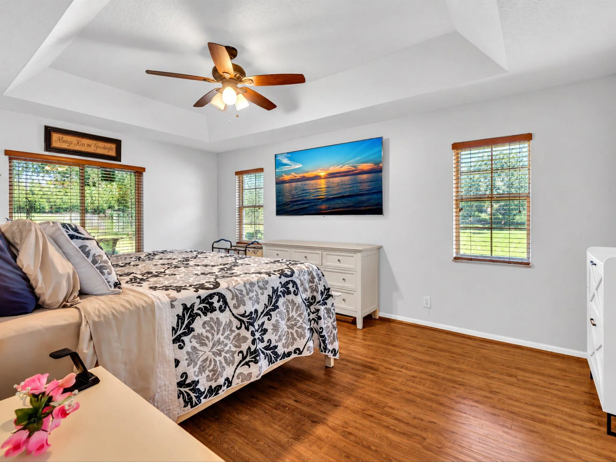 Tray ceiling with a ceiling fan, wood-finish flooring, and multiple windows with wood blinds