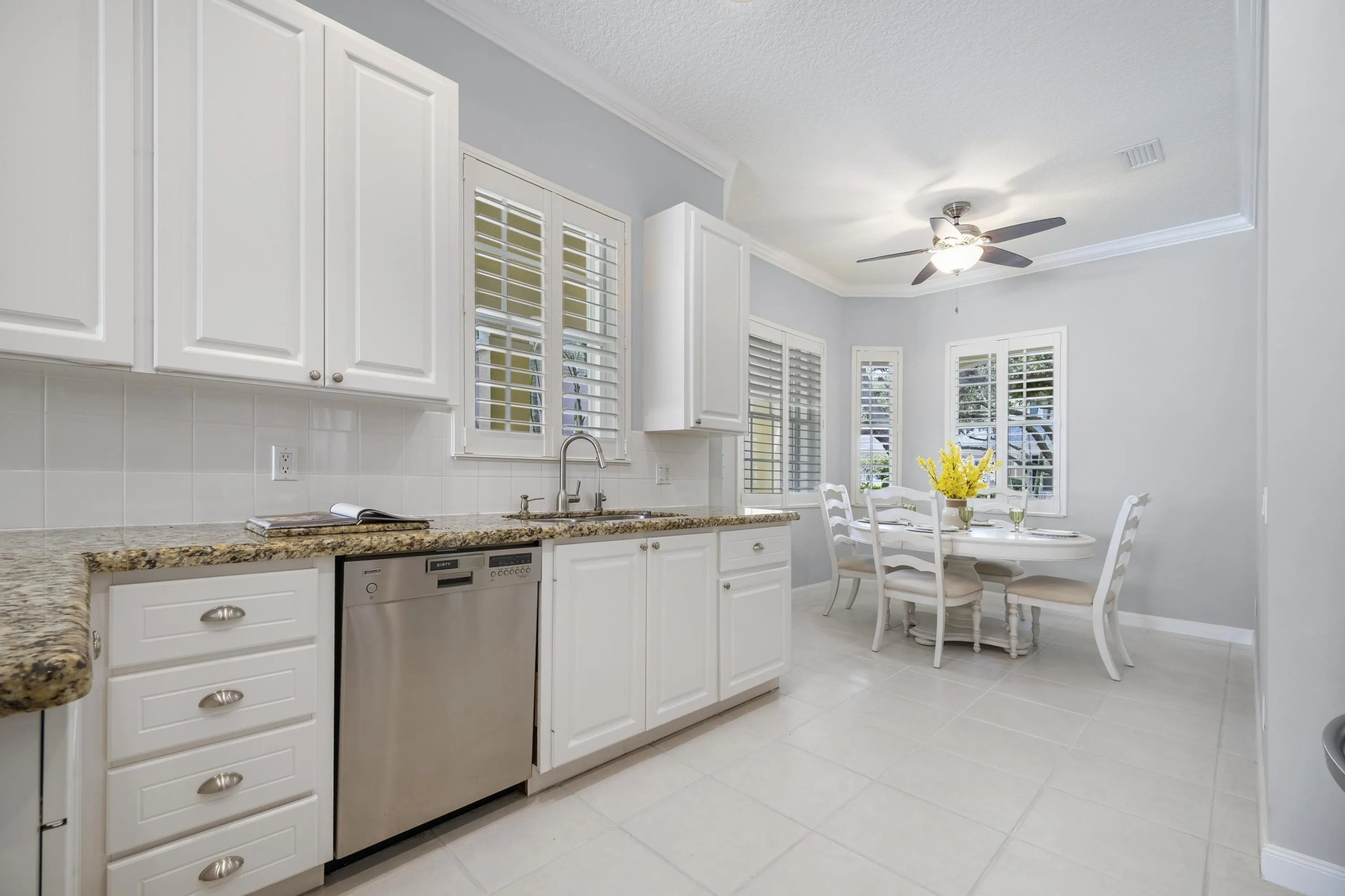 Double sink beneath plantation shutters, beside a window-lined breakfast nook with plenty of natural light