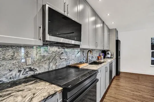 Kitchen featuring light gray cabinetry, built-in stainless steel microwave, stone countertops, stone backsplash, and wood-finish flooring