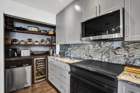 Integrated stainless steel microwave, gray cabinetry with modern hardware, stone countertops with matching full-height backsplash, wood-finish flooring, and built-in wine cooler