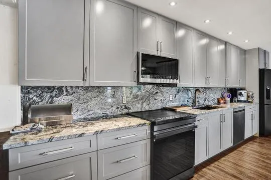 Kitchen featuring light gray shaker-style cabinetry, patterned stone countertops, a full-height patterned stone backsplash, and black appliances