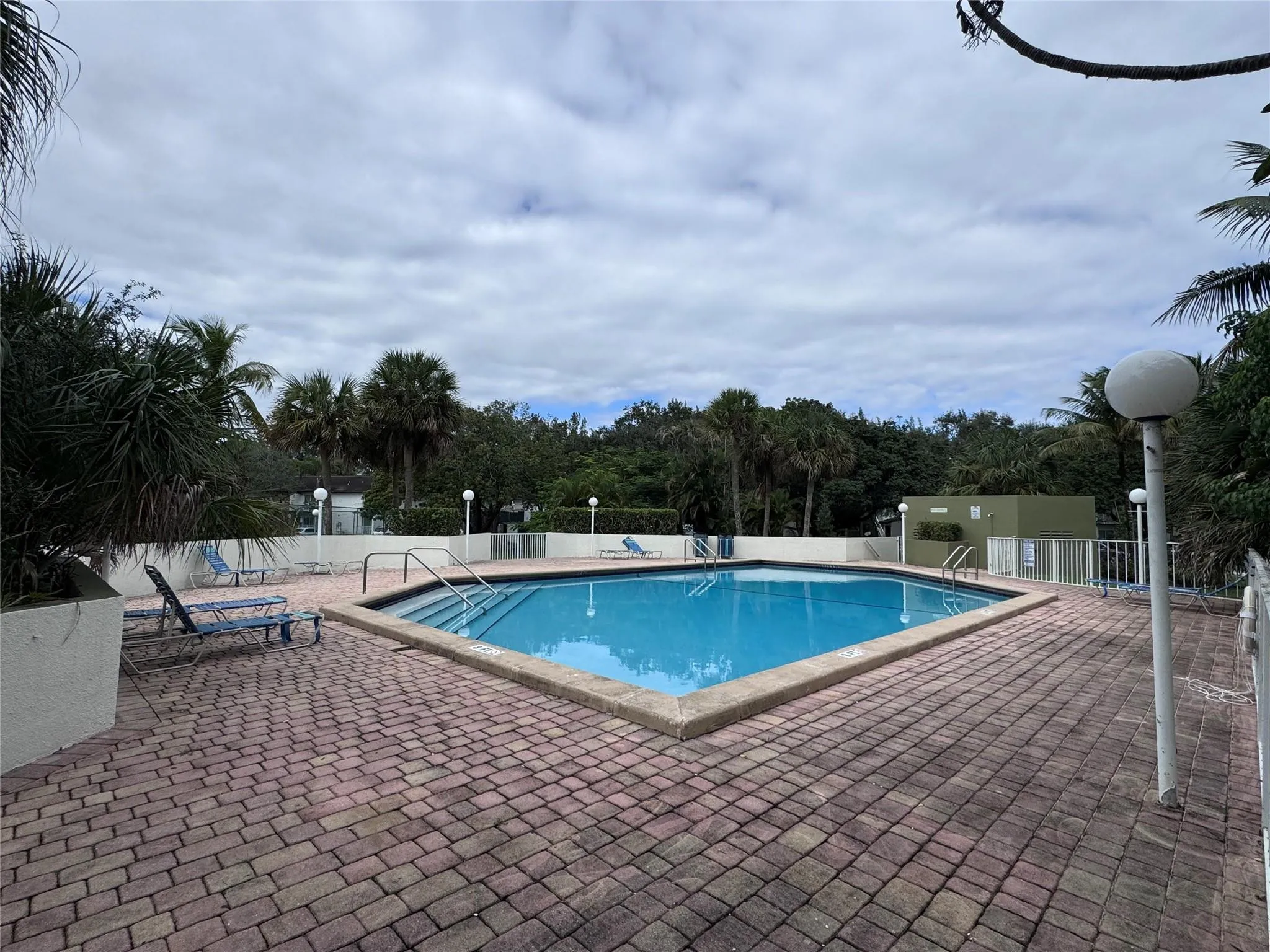 Diamond-shaped swimming pool with a paved brick deck, surrounded by landscaping, and featuring a perimeter fence