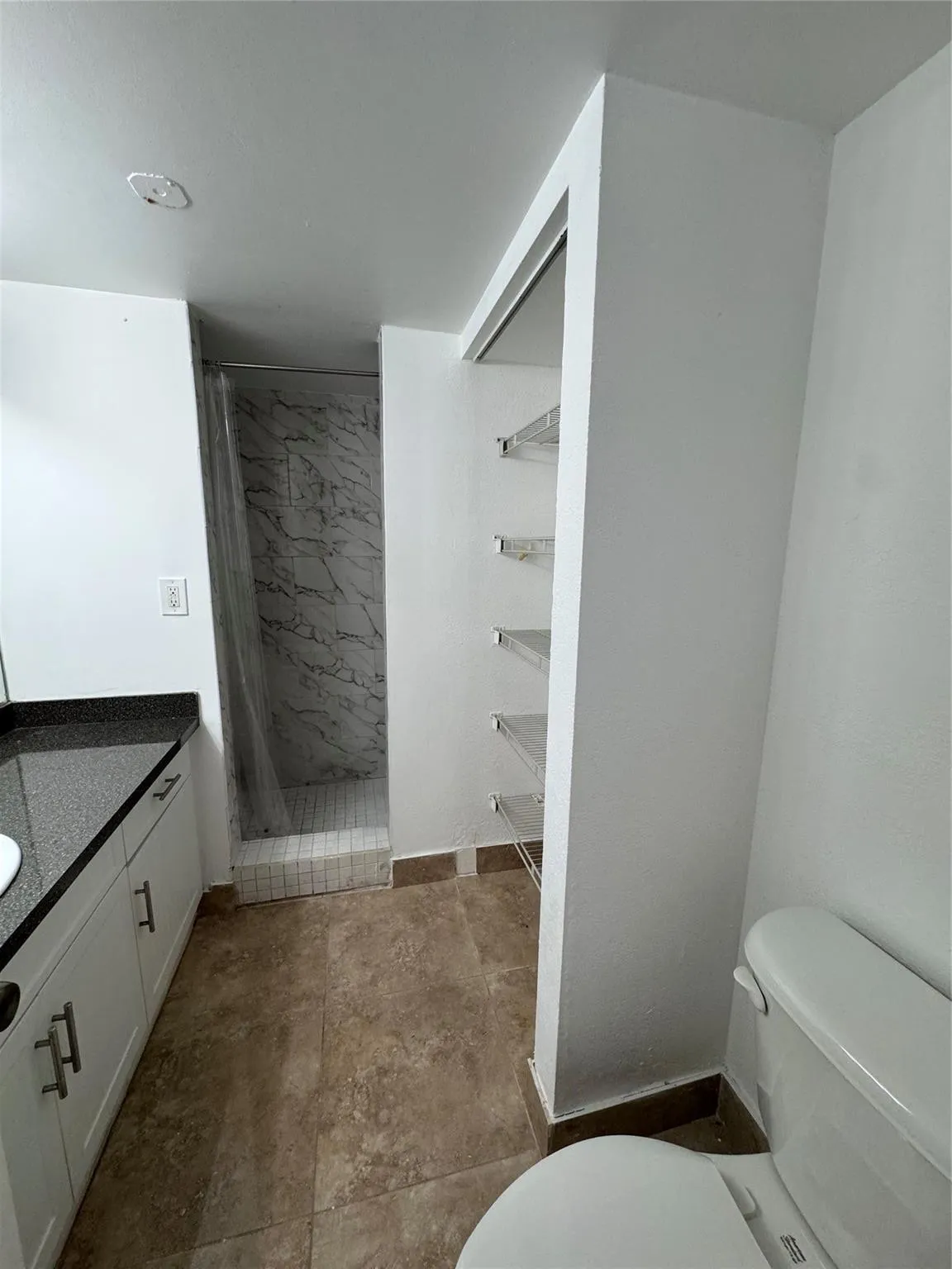 White vanity with dark stone countertop, built-in shelving, tiled flooring, and a shower with a clear enclosure