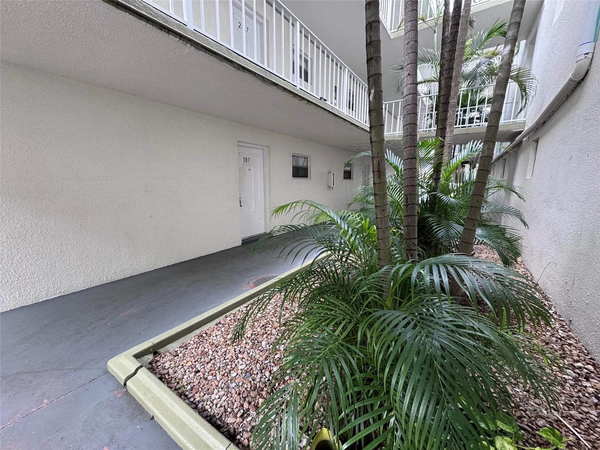 Exterior corridor with textured white walls, a gray concrete walkway, and a landscaped planter featuring palm trees and gravel