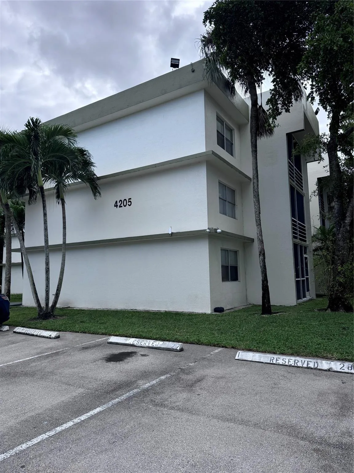 Multi-story building featuring a flat roofline, perimeter landscaping, and an adjacent asphalt parking area with marked spaces