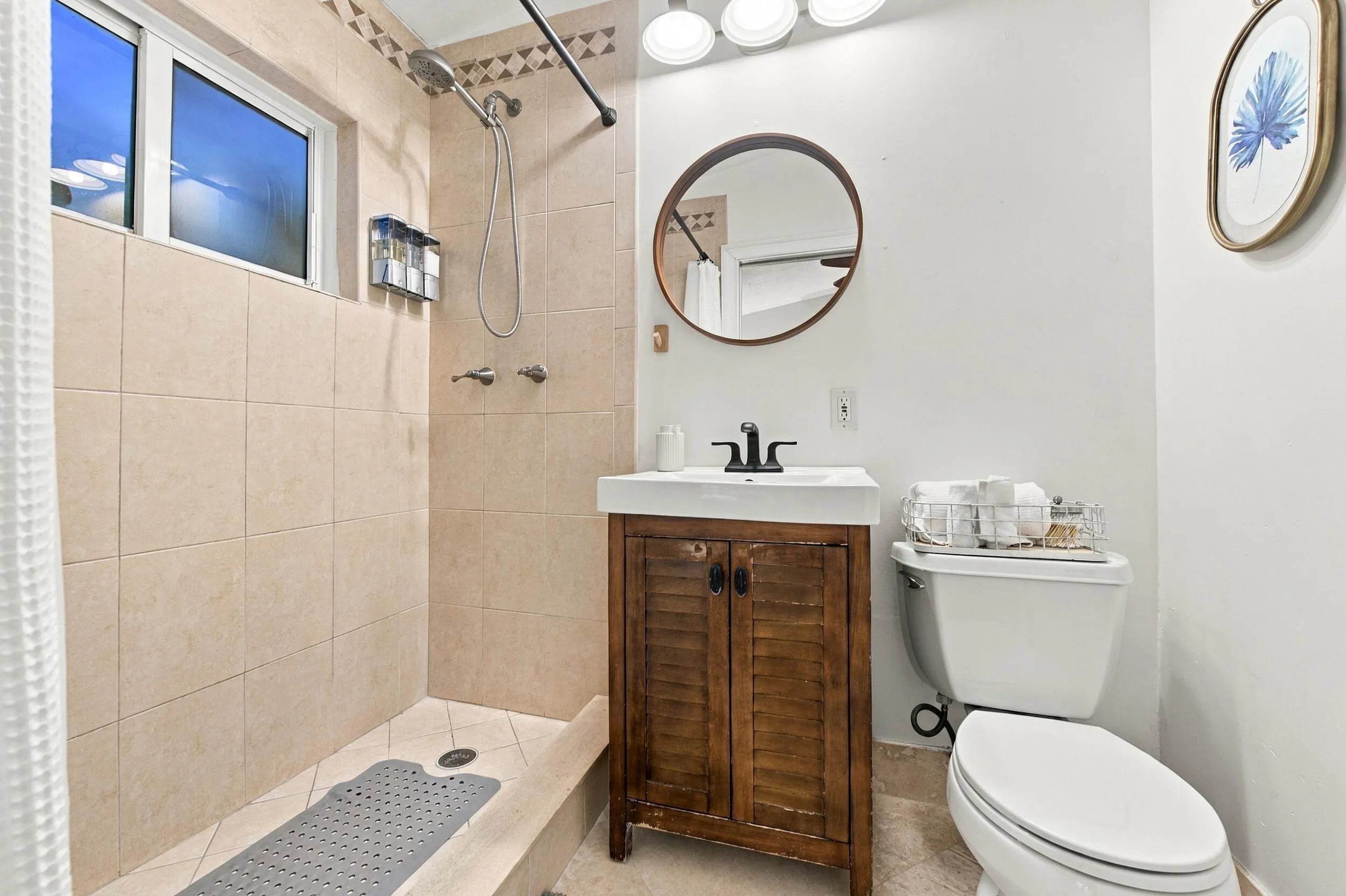 Walk-in shower with tiled walls, natural light window, dark wood-finish vanity with countertop sink, and a circular wall-mounted mirror