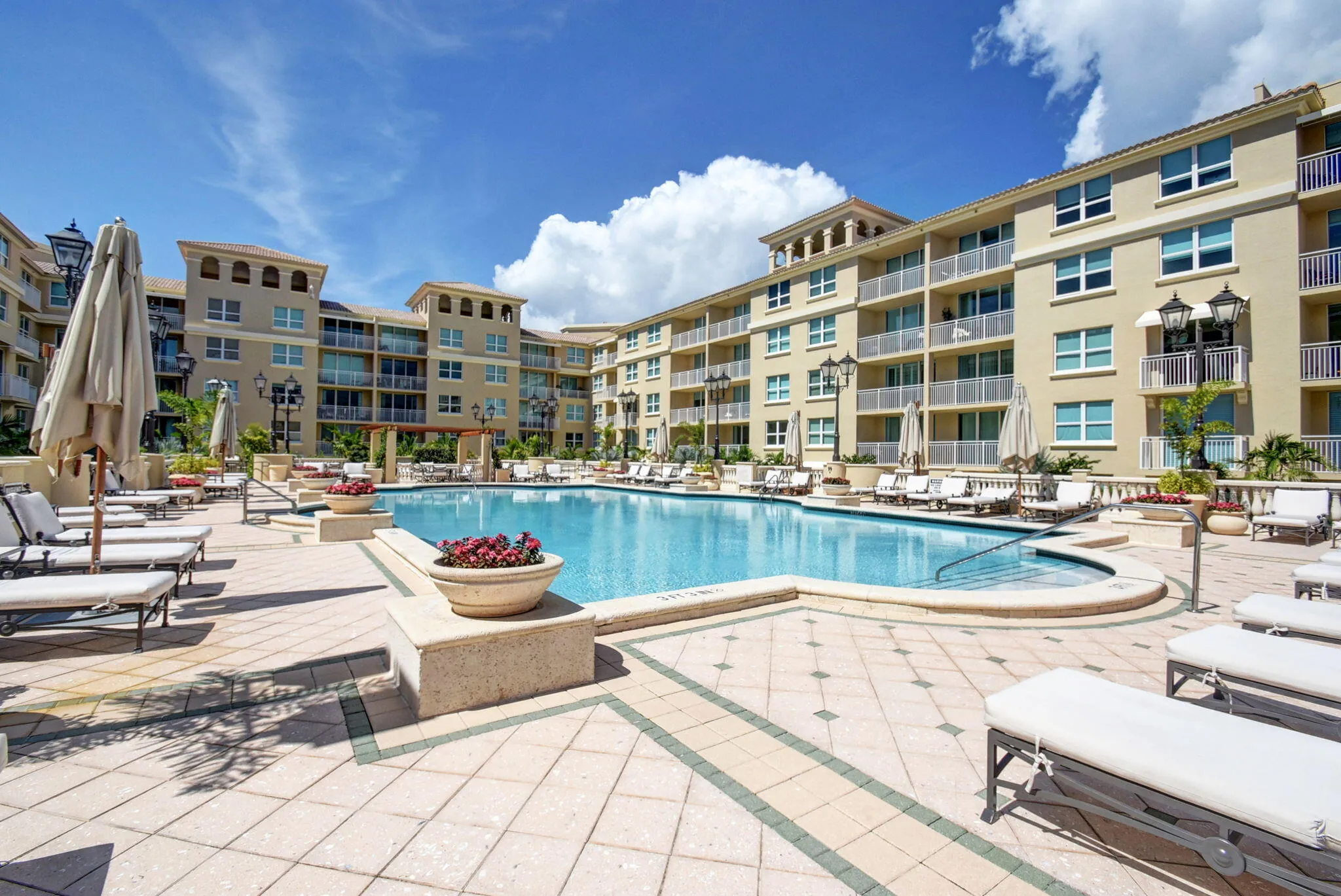 Resort-style pool with integrated spa, surrounded by extensive paver decking, decorative planters, and multiple story residential buildings with balconies