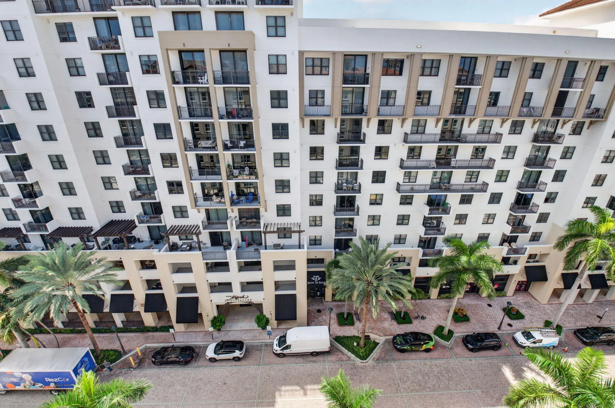Multi-story building featuring multiple private balconies with black railings, a street-level entrance with an awning, and palm trees lining the street