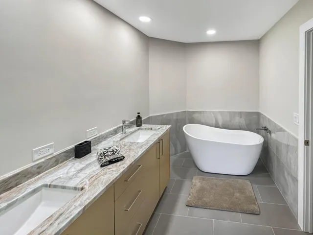 Dual vanity with integrated sinks, stone countertop, freestanding soaking tub, and tile flooring