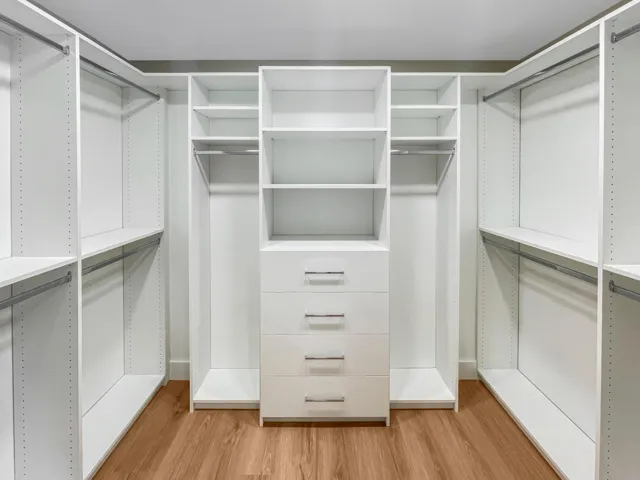 Built-in white custom closet system featuring multiple shelves, hanging rods, and a central dresser with four drawers, all set on wood-finish flooring