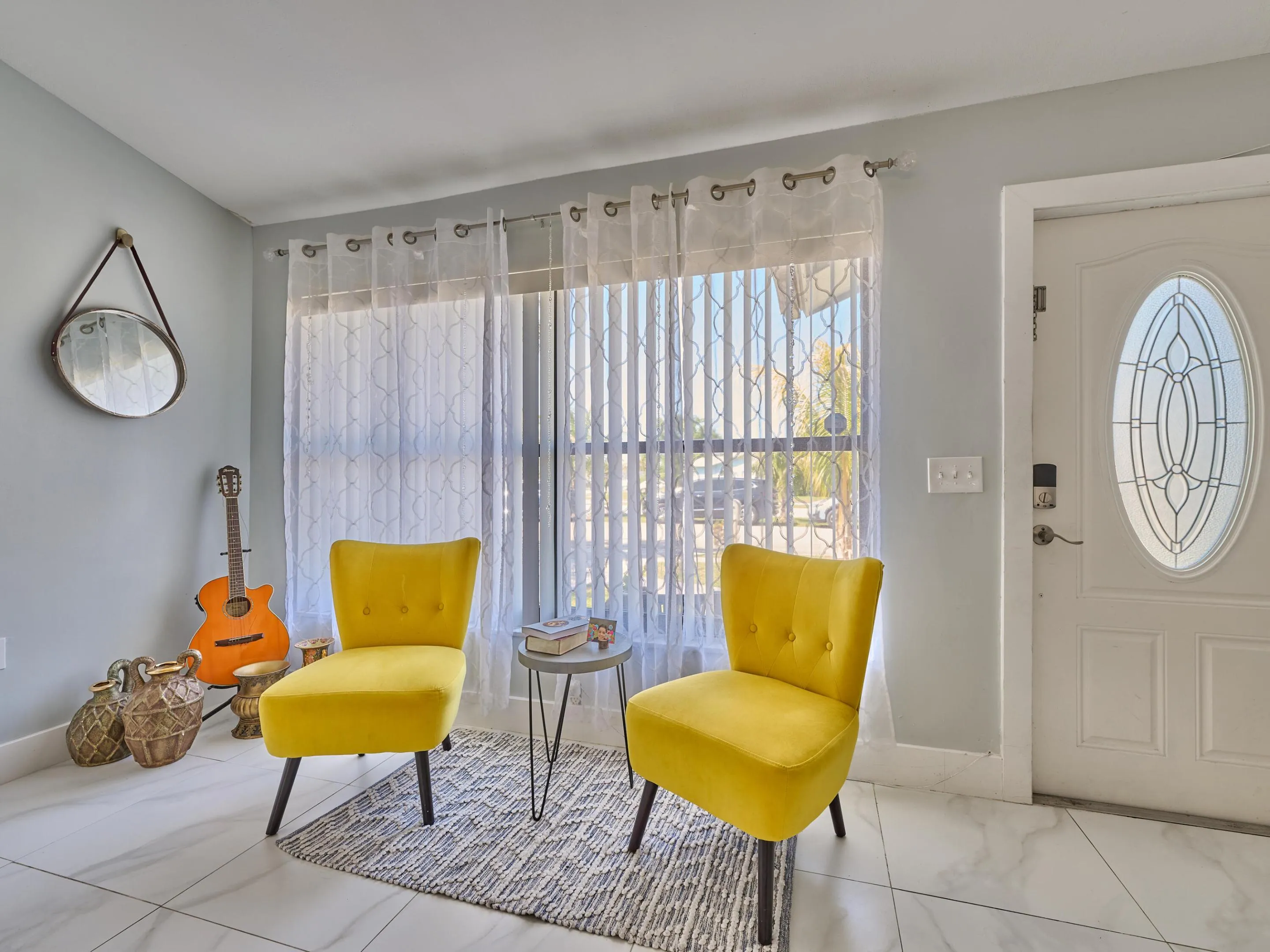 White entry door with an oval decorative glass insert, large window with sheers, and light-toned tile flooring