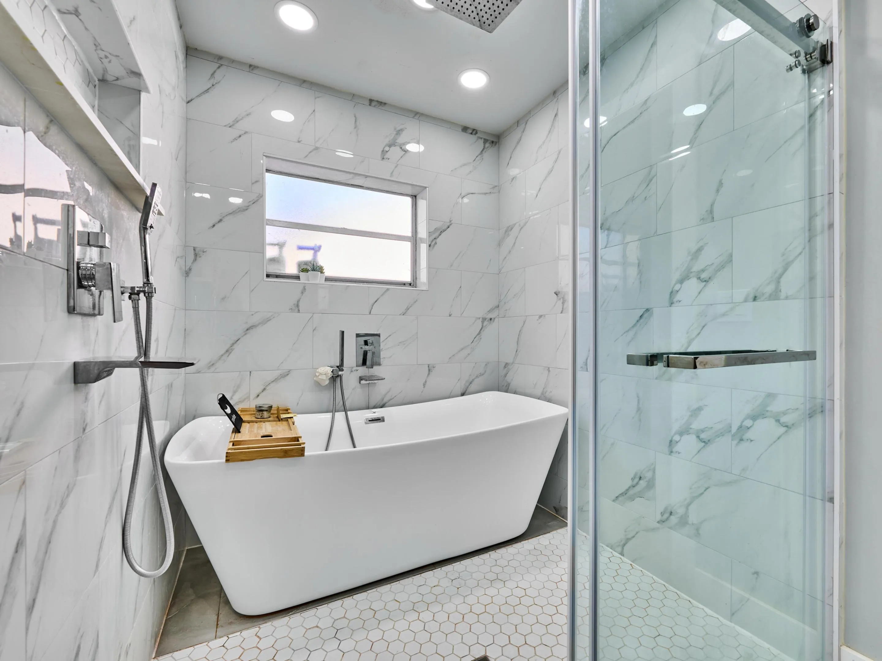 Freestanding soaking tub with floor-mounted filler, glass-enclosed shower, white tile walls with gray veining, and hexagonal floor tiling
