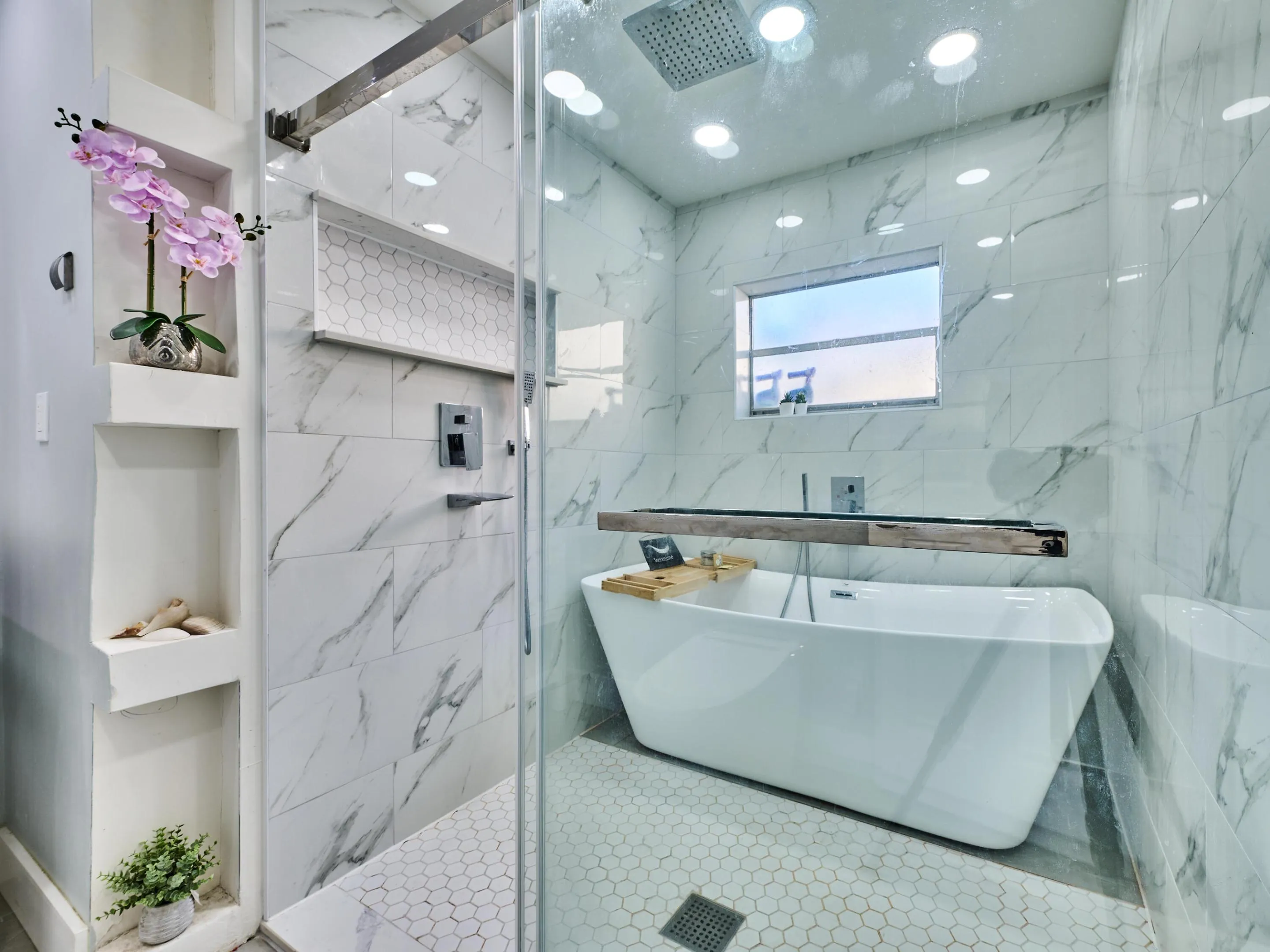 Freestanding soaking tub, floor-to-ceiling marble-look tile, glass-enclosed shower, rain shower head, and recessed wall shelving