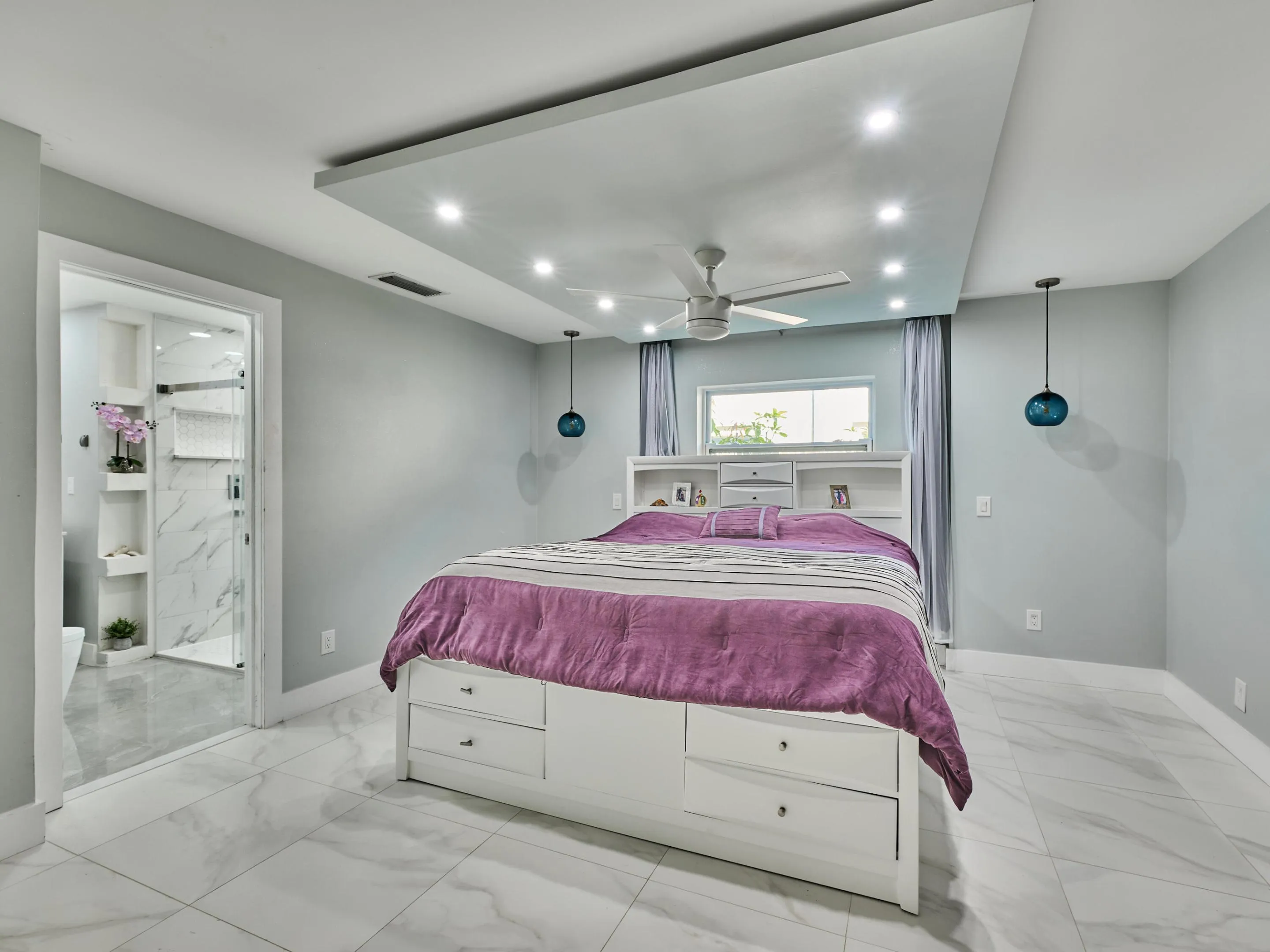 Recessed lighting in a tray ceiling, ceiling fan, large format marble-look tile flooring, and an en-suite bathroom with a glass-enclosed shower