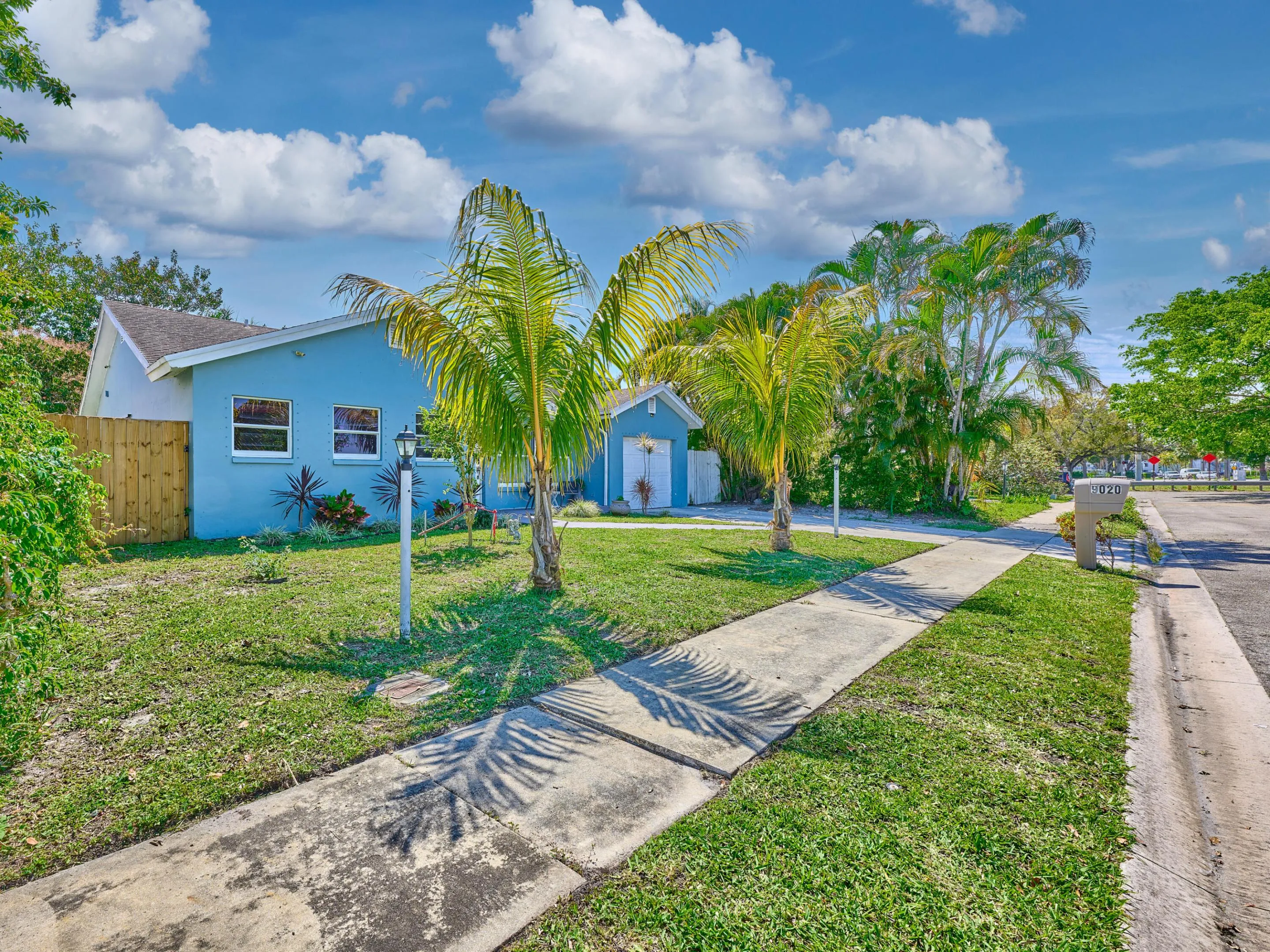 Blue exterior, white-framed windows, wood fencing, mature palm trees, and sidewalk access