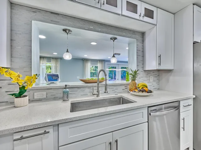 White shaker-style cabinetry with dark pulls, stone-finish countertops, stainless steel sink with gooseneck faucet, full-size stainless steel dishwasher, and a light-toned tile backsplash