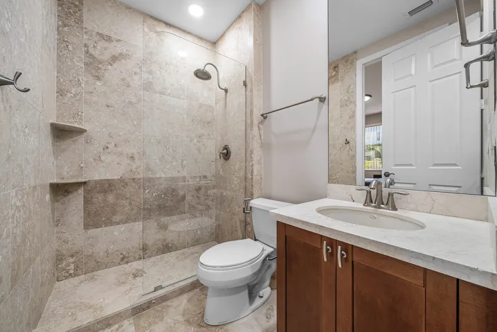 Full vanity with wood-finish cabinetry, under-mount sink, and dual-handle faucet with travertine marble floor to ceiling and modern frameless glass shower door