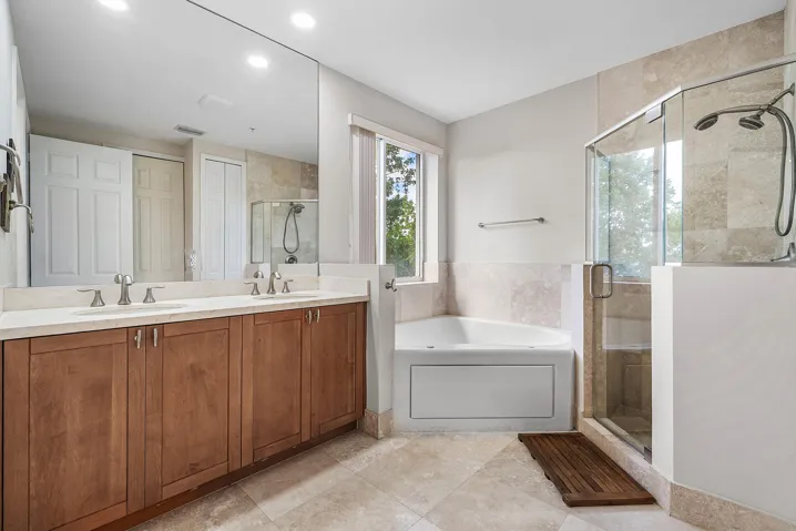 Dual vanity with wood cabinetry and undermount sinks, corner soaking tub, glass-enclosed shower with tiled walls, and large format marble flooring