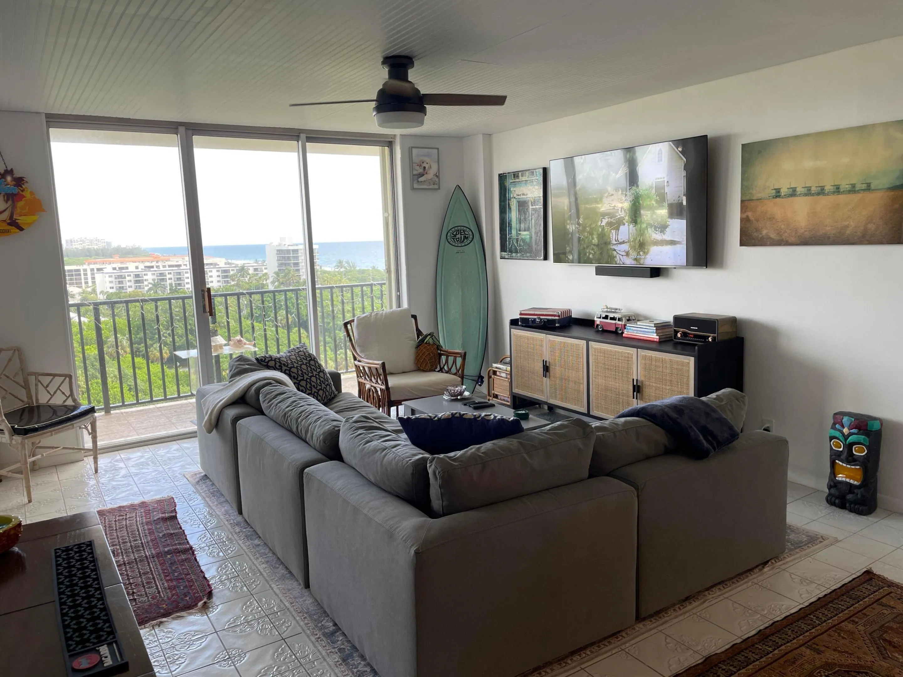 Sliding glass doors opening to a balcony, white tongue-and-groove ceiling, ceiling fan with integrated lighting, wall-mounted media console, and white tile flooring