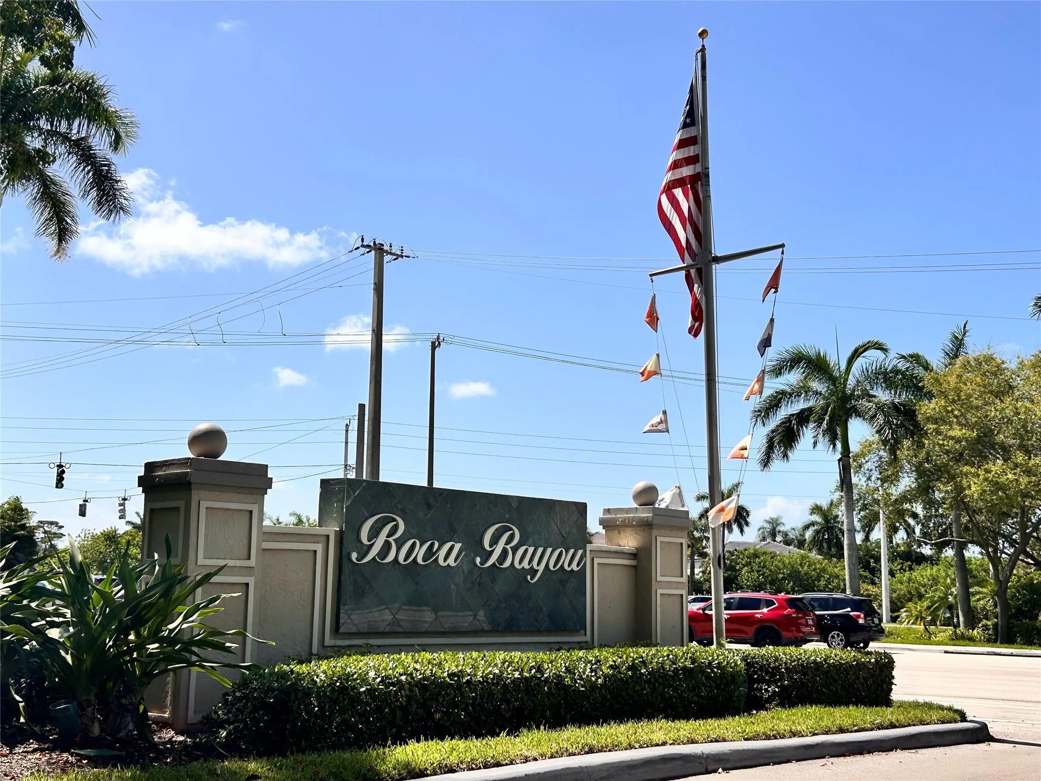 Gated community entrance with a prominent stone sign, mature landscaping, and a flag pole