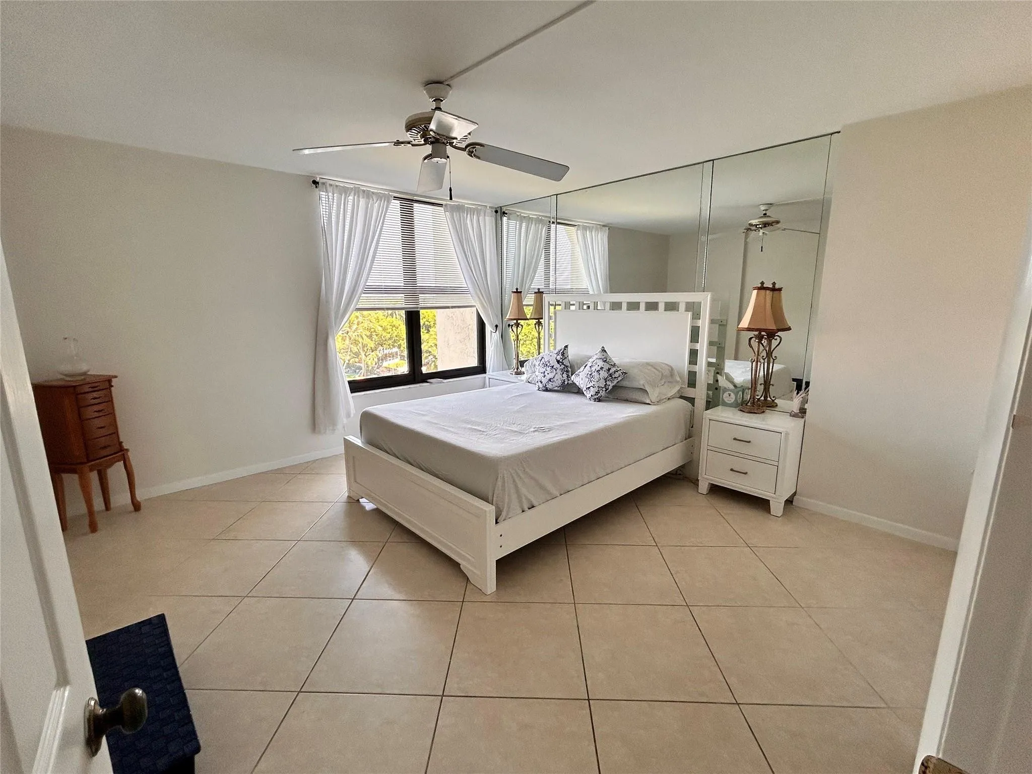Large format tile flooring, expansive window, ceiling fan with integrated lighting, full-wall mirrored accent, and crisp white walls