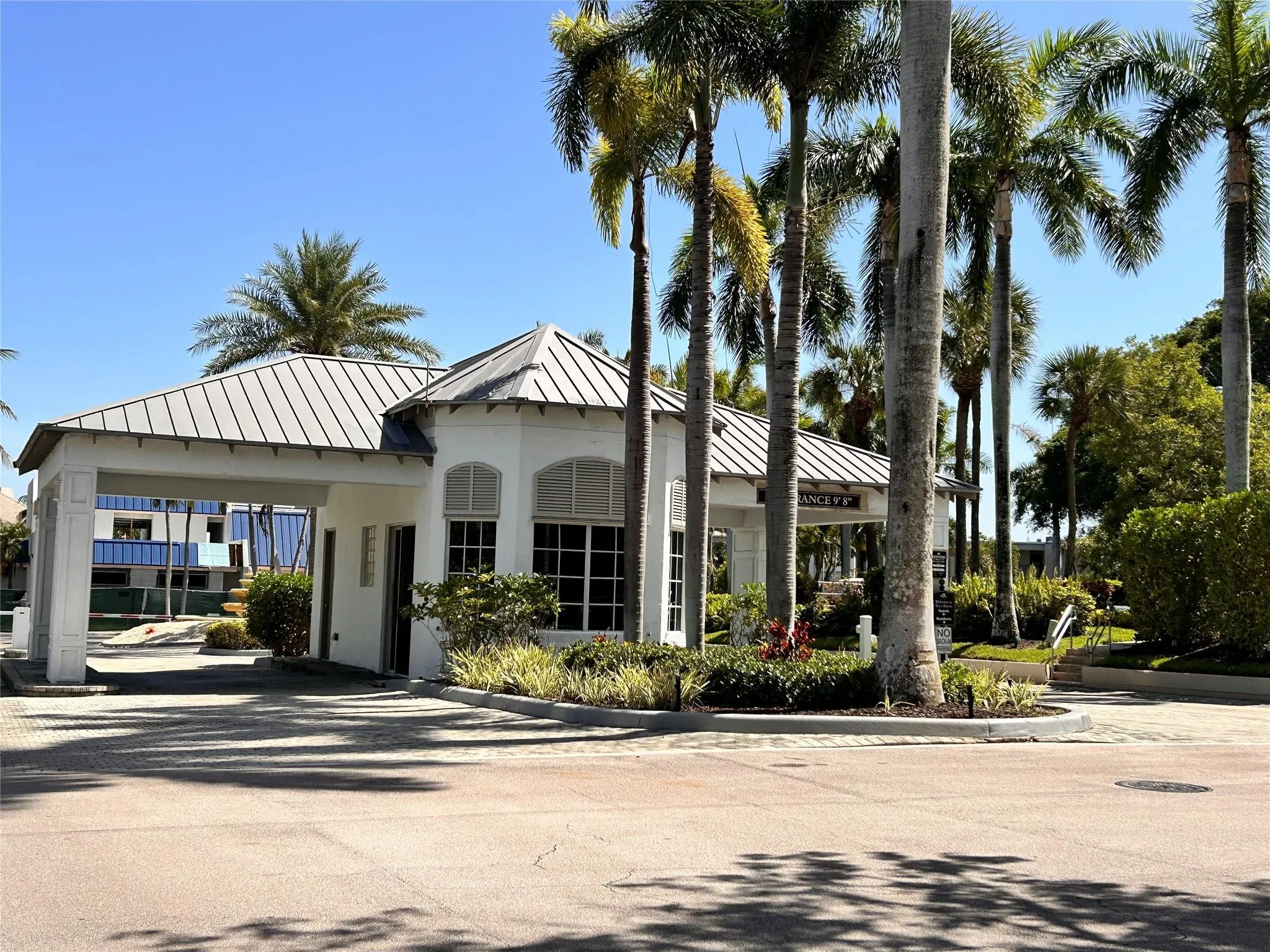 White stucco building with a metal seam roof, featuring multiple tall palm trees and paver driveway