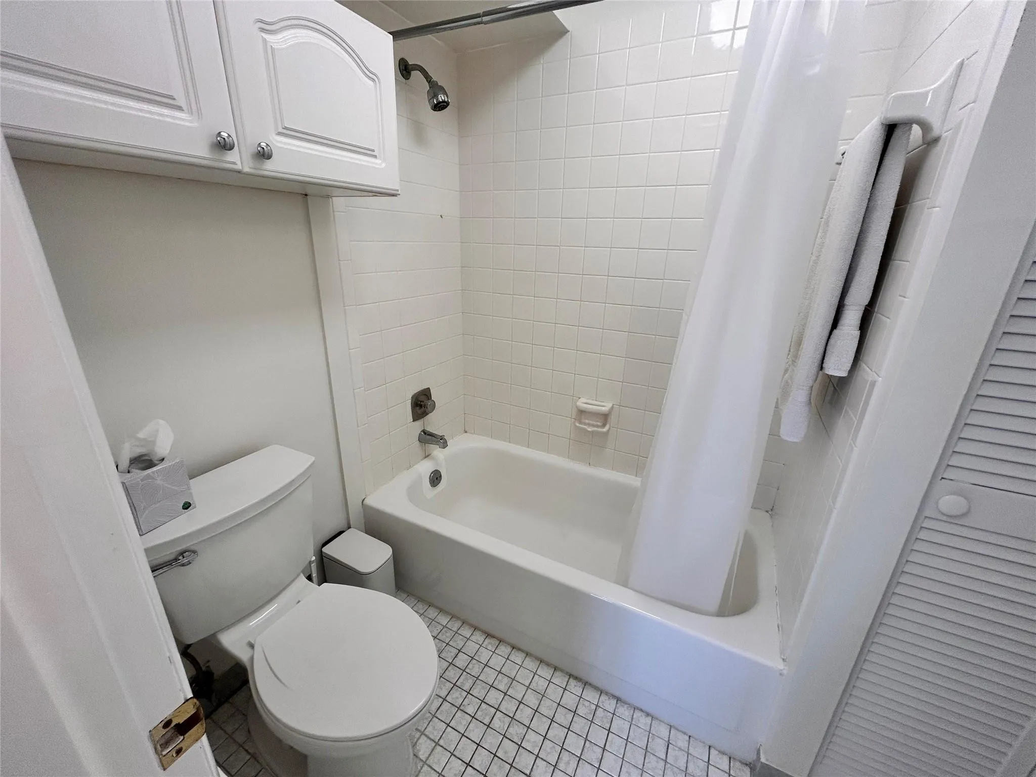White ceramic tile flooring, bathtub with an integrated shower, wall-mounted showerhead, and white cabinetry with arched panels