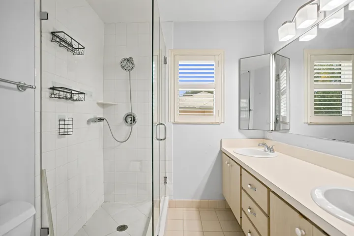 Glass-enclosed shower with white tiled walls, dual vanity with undermount sinks, wood-finish cabinetry, and shuttered windows