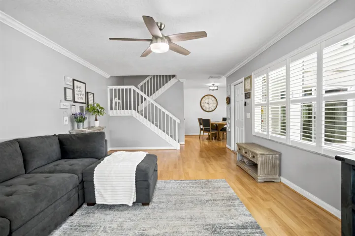 Wood-finish flooring, staircase with white risers and railings, ceiling fan with integrated lighting, multiple windows with white plantation shutters, and crown molding