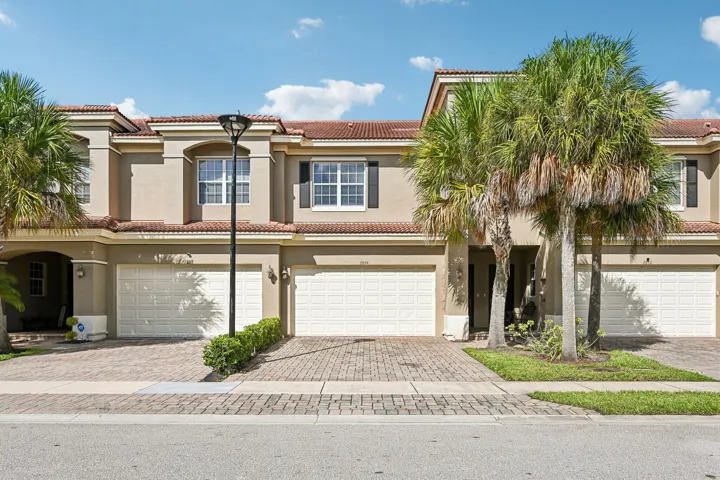 Stucco exterior with tile roof, paver driveway, attached two-car garage, and established palm trees