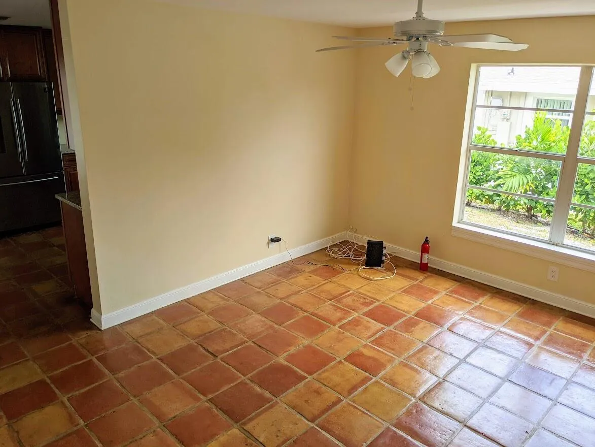 Terracotta-style floor tiles, ceiling fan with integrated lighting, kitchen featuring stainless steel refrigerator and wood-finish cabinetry, and a large window providing ample natural light