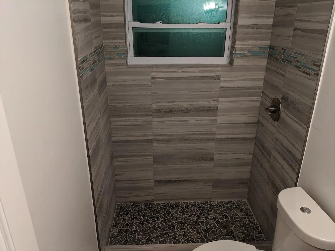 Tiled shower enclosure featuring horizontal gray tile, pebble-finish flooring, accent mosaic tile band, and a window