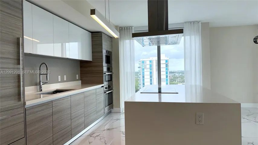 Contemporary kitchen featuring a stone-topped island with an integrated cooktop, two-toned cabinetry, stainless steel wall-mounted oven and microwave, stone-finish flooring, and a stainless steel range hood