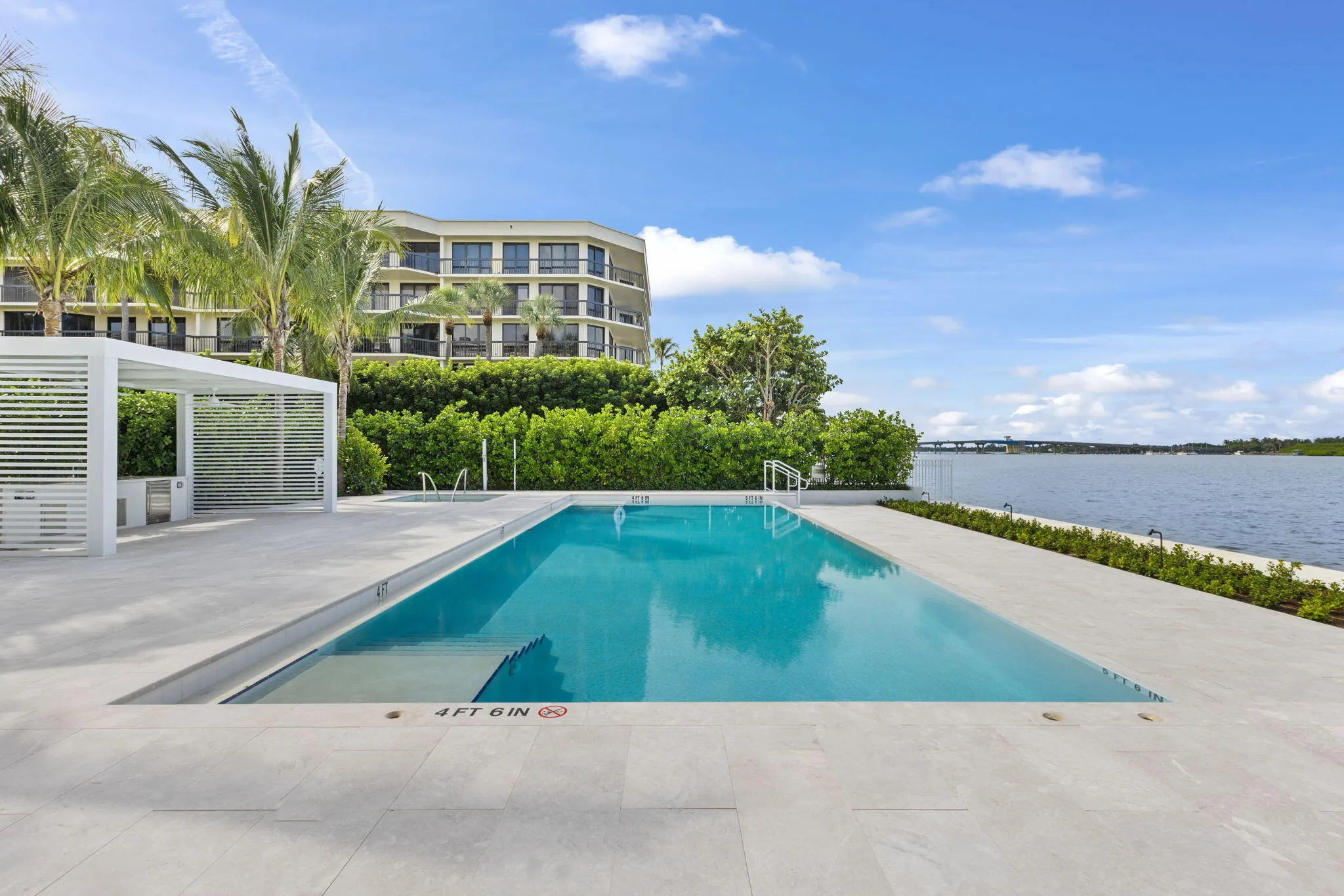In-ground swimming pool, white pergola, hardscaping with light-toned pavers, mature landscaping, and waterfront access