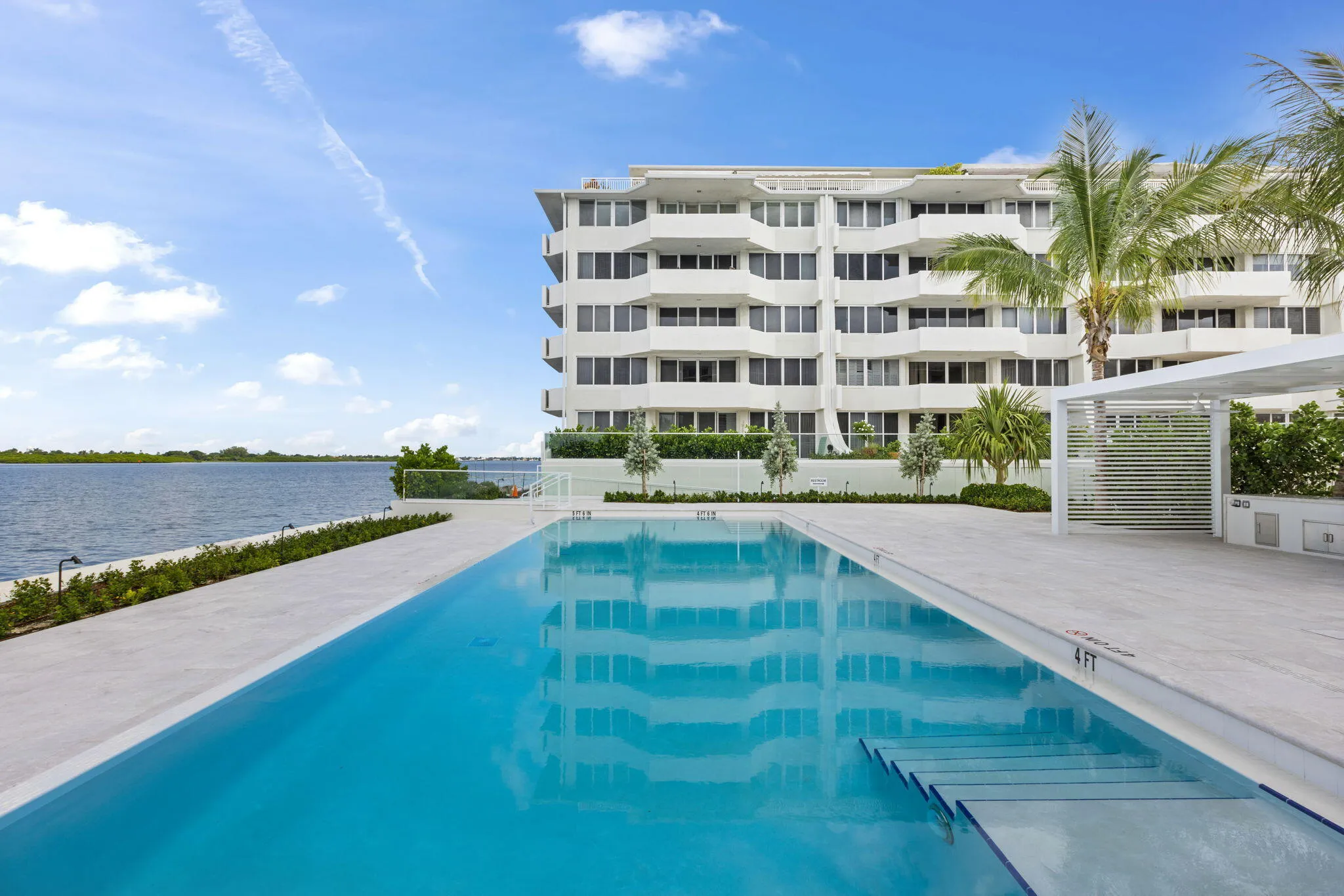 Modern high-rise residential building with multiple balconies, expansive outdoor swimming pool with submerged steps, white hardscaping, and a contemporary pergola structure
