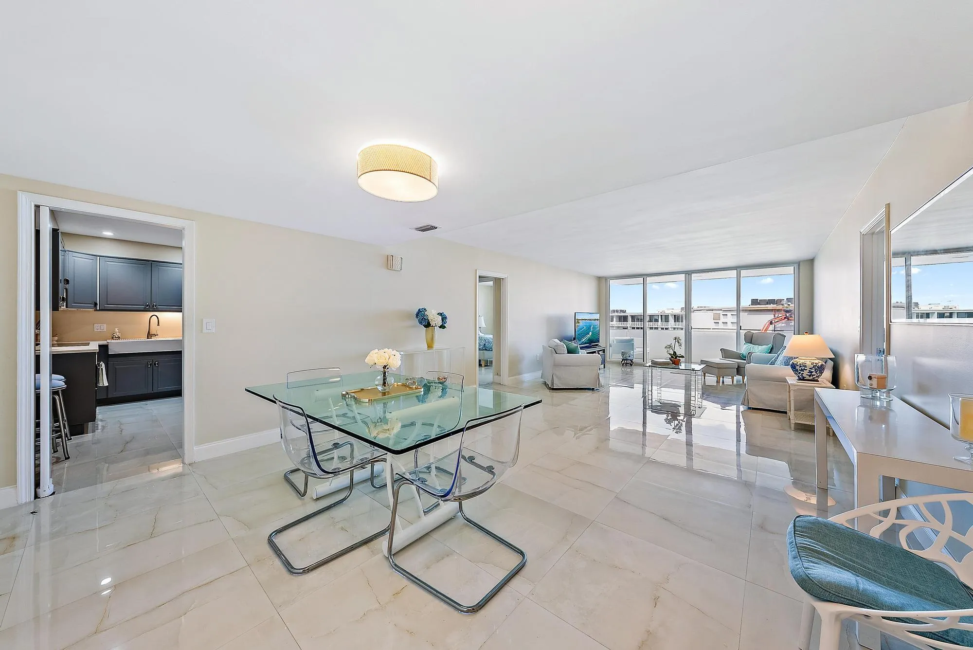 Expansive open-concept living area showcasing polished light-stone flooring, floor-to-ceiling windows with panoramic outlooks, recessed ceiling lighting, and a modern glass-top dining table with sculptural base