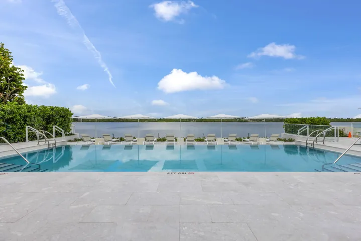 Expansive outdoor pool with integrated steps, surrounded by a light-toned paved deck, and featuring a clear glass safety barrier