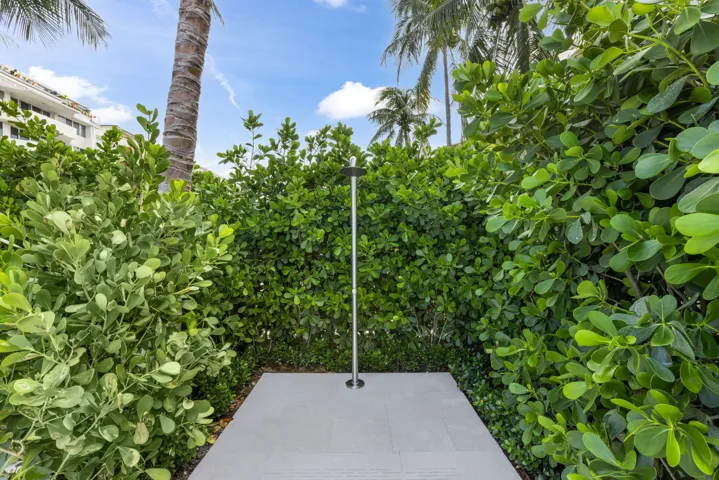 Outdoor shower with a stainless steel fixture, surrounded by dense privacy hedging and mature palm trees