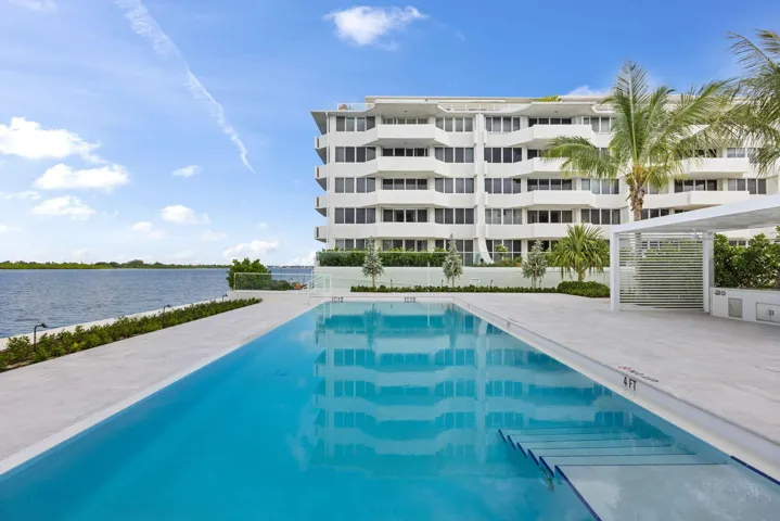 Modern high-rise residential building with multiple balconies, expansive outdoor swimming pool with submerged steps, white hardscaping, and a contemporary pergola structure