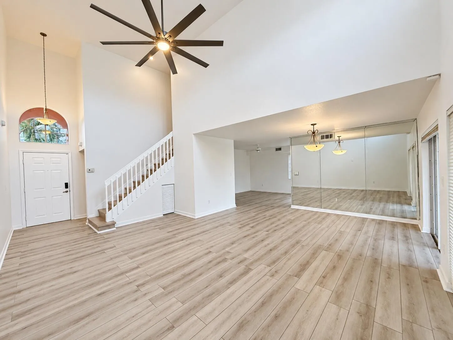 Vaulted ceiling with a large ceiling fan, wood-finish flooring, staircase with white railing, wall-mounted mirrors, and an arched transom window above the entry door