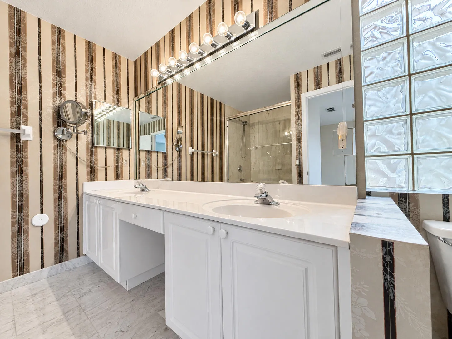 Dual vanity with integrated sinks, white cabinetry, a large mirror, and a glass block window