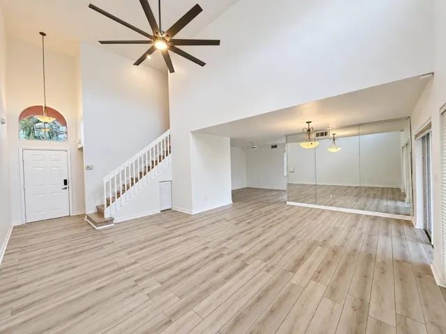 Vaulted ceiling with a large ceiling fan, wood-finish flooring, staircase with white railing, wall-mounted mirrors, and an arched transom window above the entry door