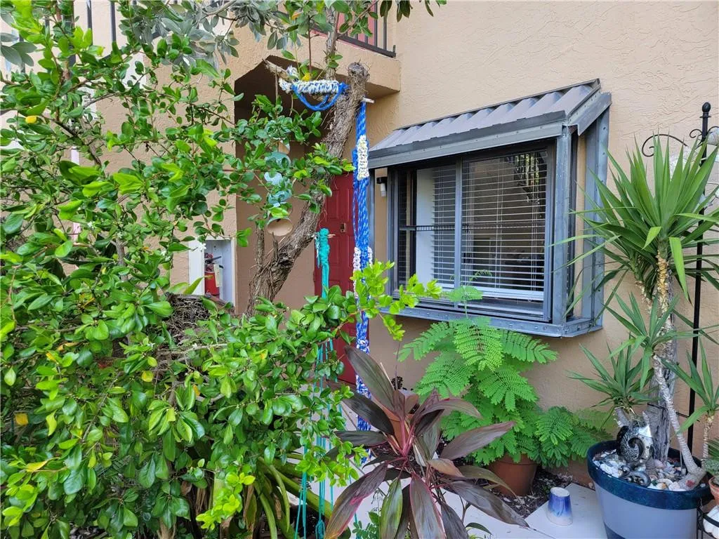 Stucco exterior with an angled window awning, framed by lush foliage and mature plantings